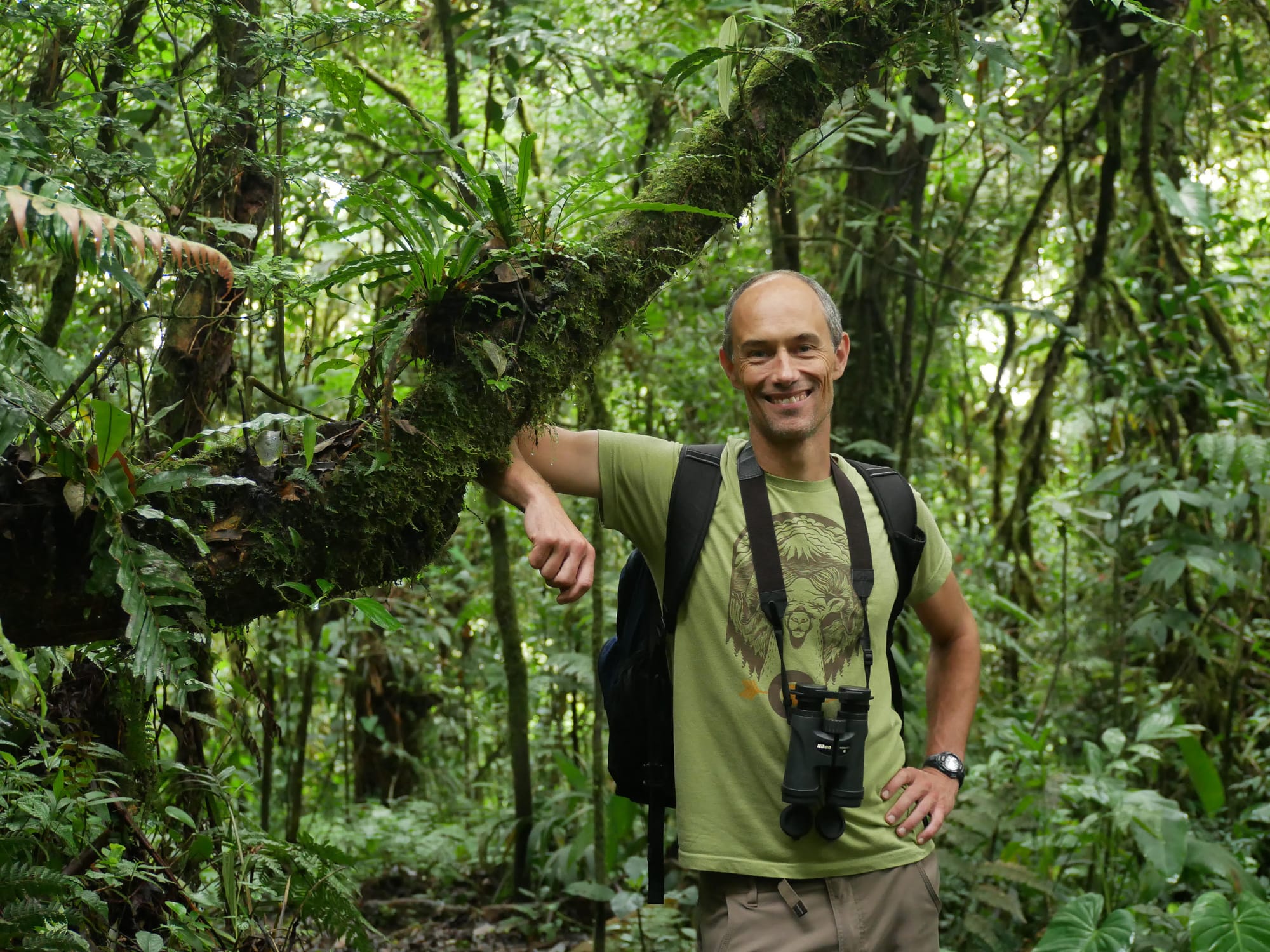 A man in his 40s or early 50s with pale skin and short gray hair leans against a tree and looks directly into the camera. He's wearing a green shirt, tan pants, a black backpack, and binoculars. The background is a vibrant green rainforest absolutely overflowing with plants.