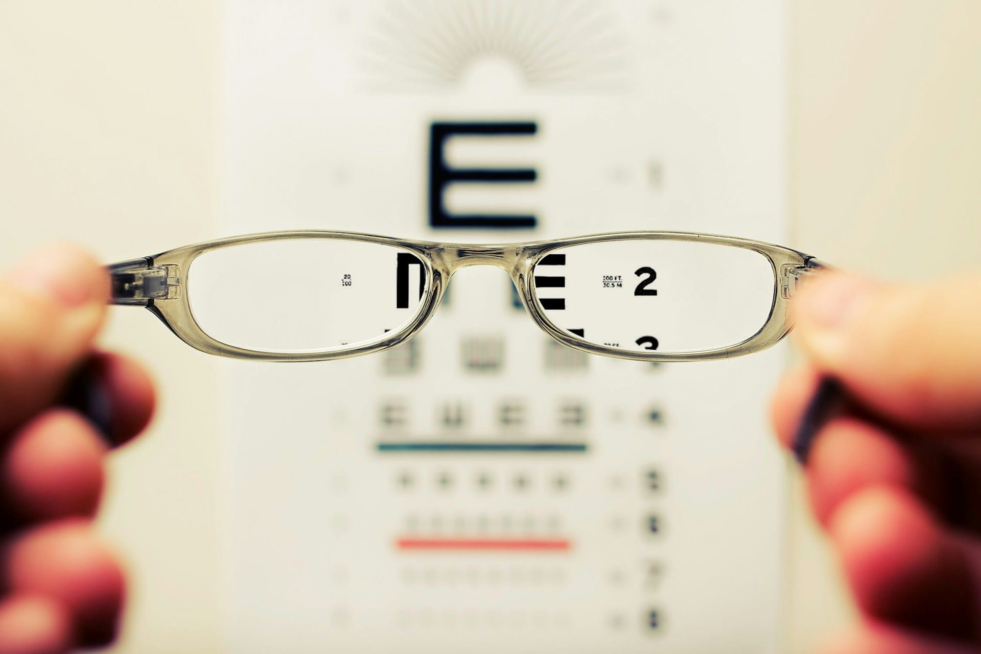A photograph of a pair of glasses being held up in front of a vision test board wiht letters and numbers. The picture is all blurry, except through the glasses. Through the lenses, you can see the letters and numbers sharply.