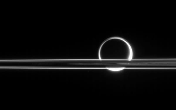 Saturn's rings slice across the moon Titan, its hazy atmosphere illuminated from behind almost like a crescent moon in this black and white photo from the Cassini spacecraft